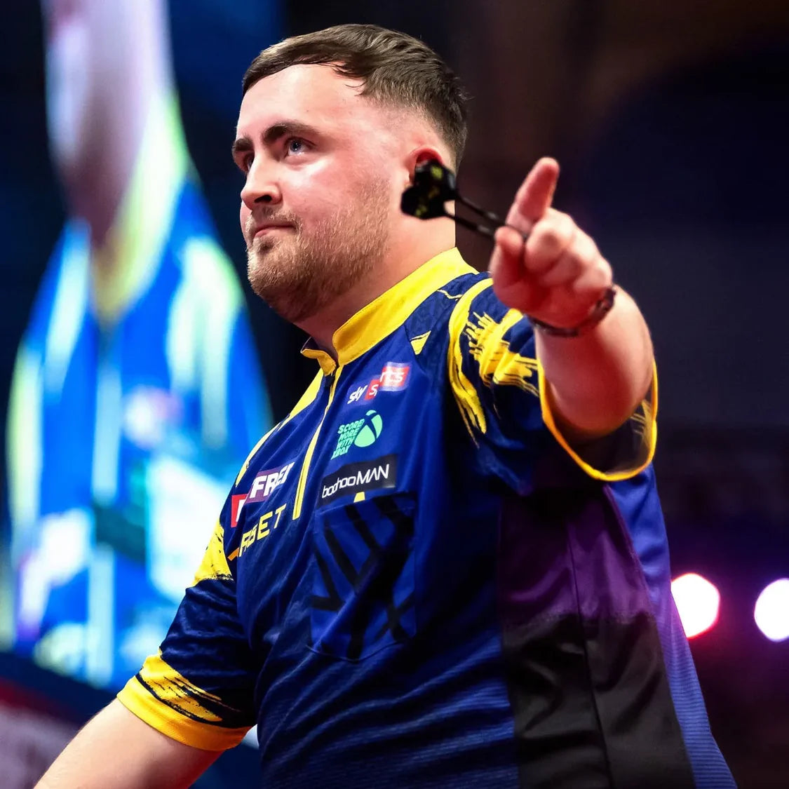 Luke Littler 2026 darts shirt in action, player on stage wearing blue and yellow shirt with sponsor logos during competitive throw