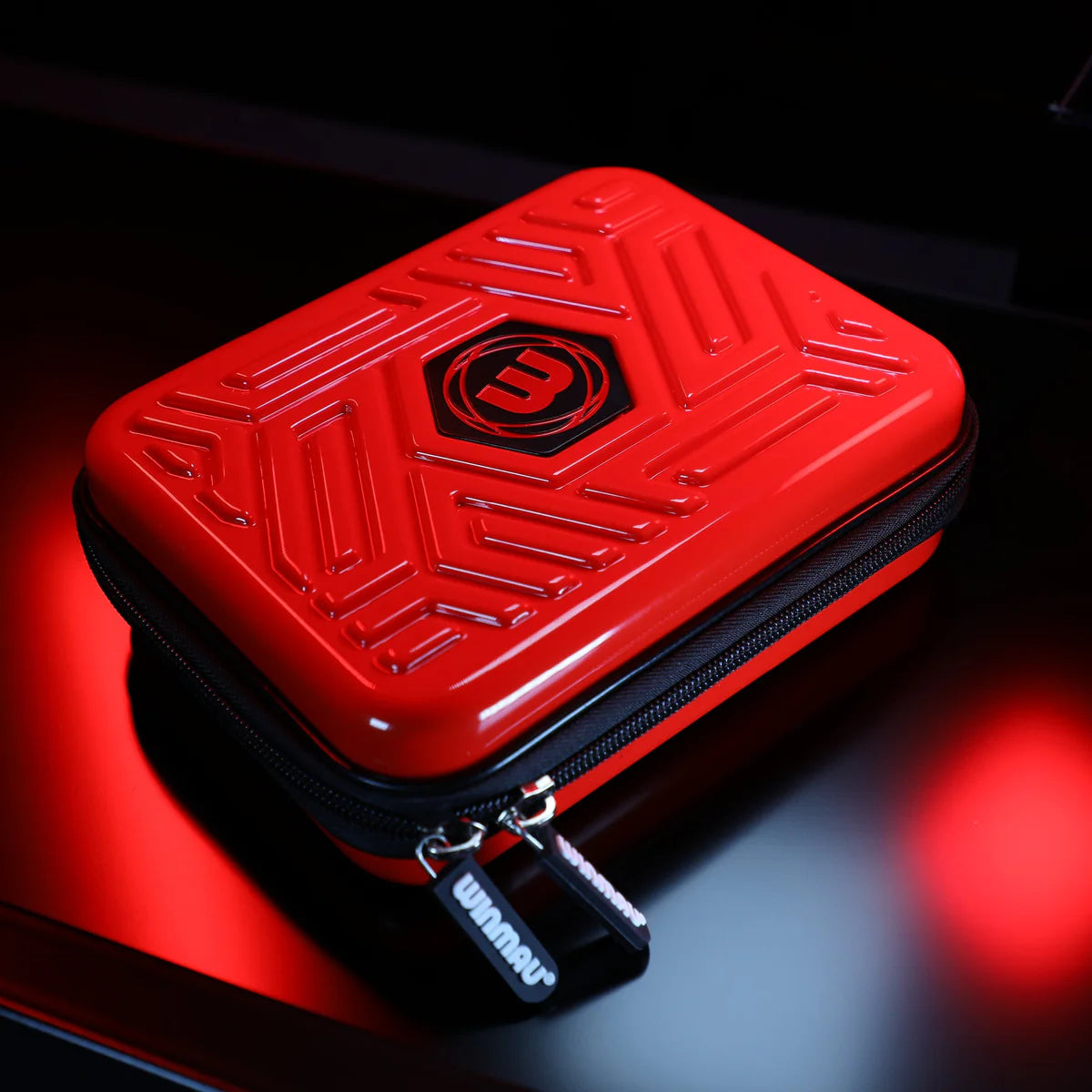 Angled view of red hard-shell darts case with raised geometric texture, black zipper, and molded logo, isolated on dark background