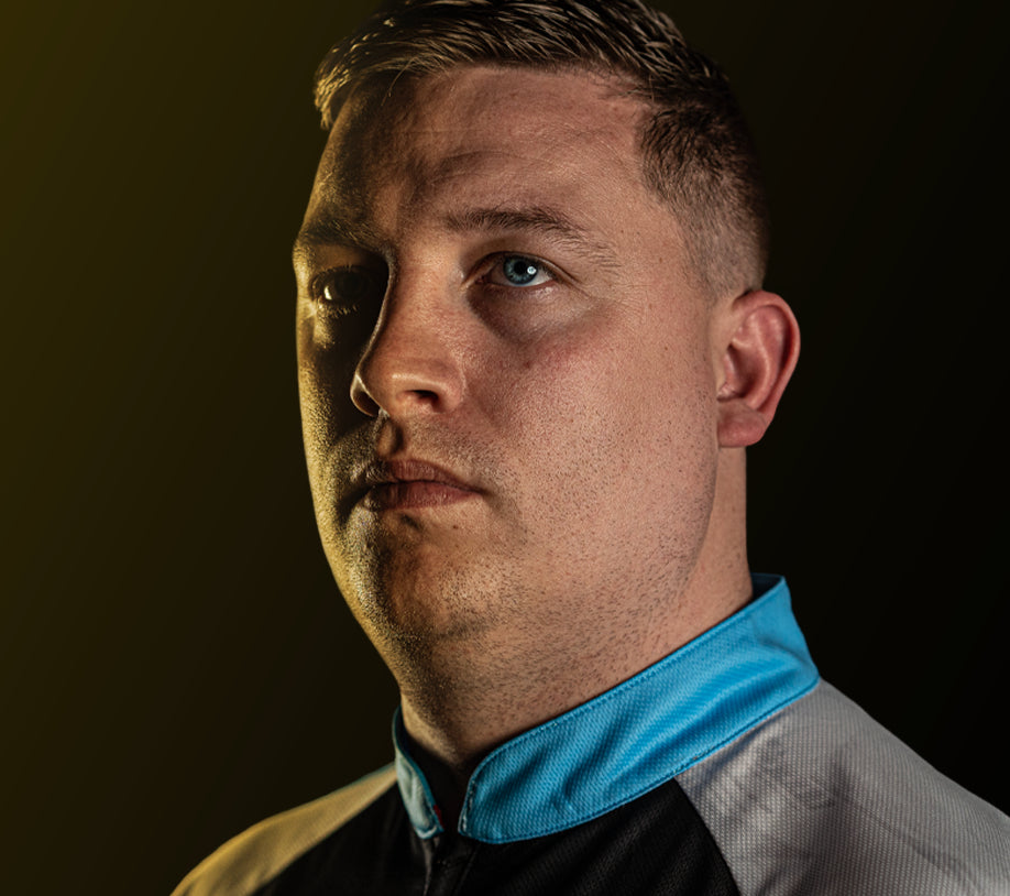 Close-up headshot of professional darts player in blue-collared shirt, focused expression, isolated on dark background
