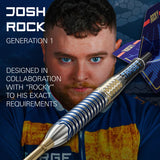 Angled close-up of Josh Rock G1 tungsten dart highlighting textured grip, blue rings, and hexagonal barrel design with player background